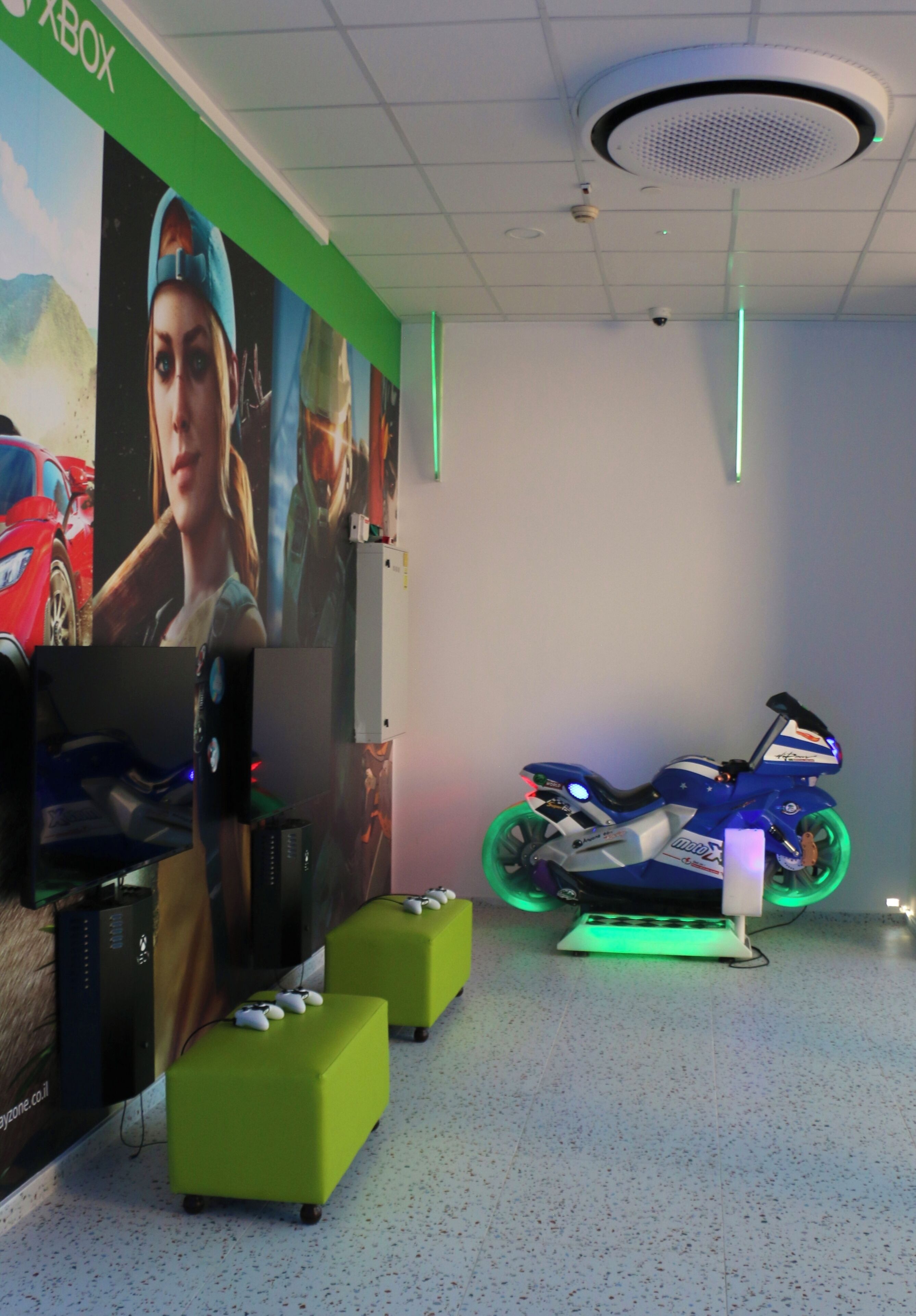 A gaming room with a large poster, a gaming console, a motorcycle, and green ottomans.