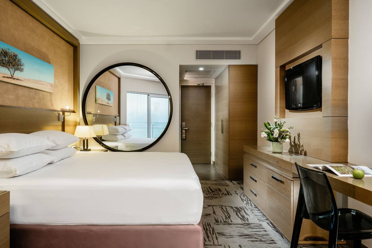 A hotel room with a bed, a desk, a chair, a TV, a mirror, and a view of the balcony.
