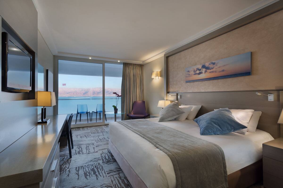 A hotel room with a large bed, a desk, a chair, a TV, and a view of the mountains.