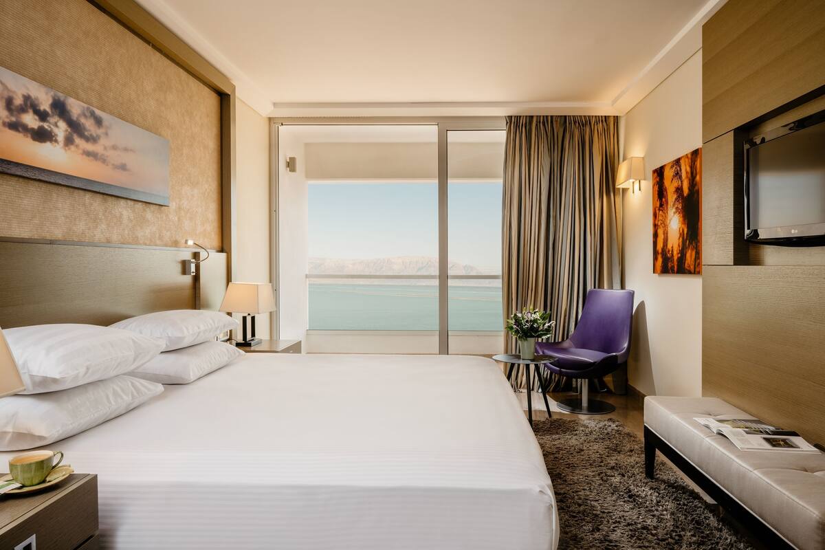 A hotel room with a large bed, a chair, a small table, a TV, and a view of the water.