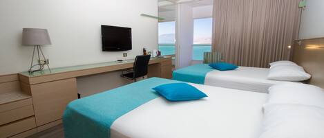 A hotel room with two beds, a desk, a television, and a view of the sea.