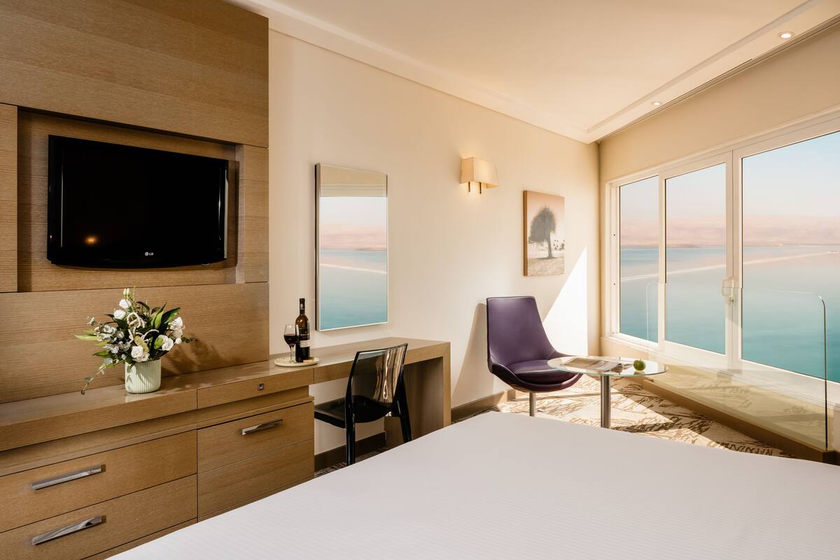 A hotel room with a large bed, a desk with a chair, a flat-screen TV mounted on the wall, a window with a view of the sea, and a vase of flowers on the dresser.