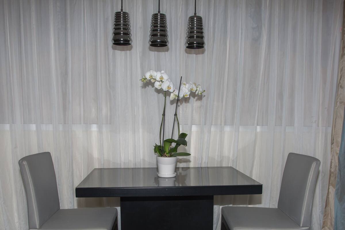 A dining area with a rectangular table, two chairs, a potted orchid, and three hanging pendant lights against a striped curtain backdrop.