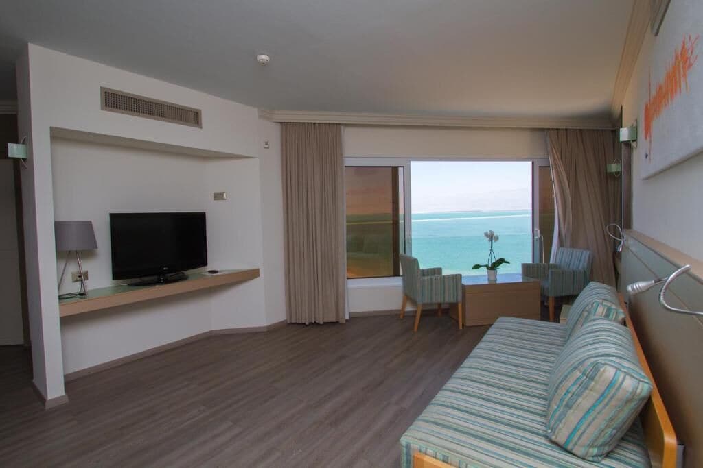 Een hotelkamer met een groot raam met uitzicht op zee, een flatscreen televisie op een houten plank, een gestreept sofa en een klein tafeltje met stoelen.