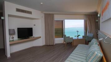 Een hotelkamer met een groot raam met uitzicht op zee, een flatscreen televisie op een houten plank, een gestreept sofa en een klein tafeltje met stoelen.