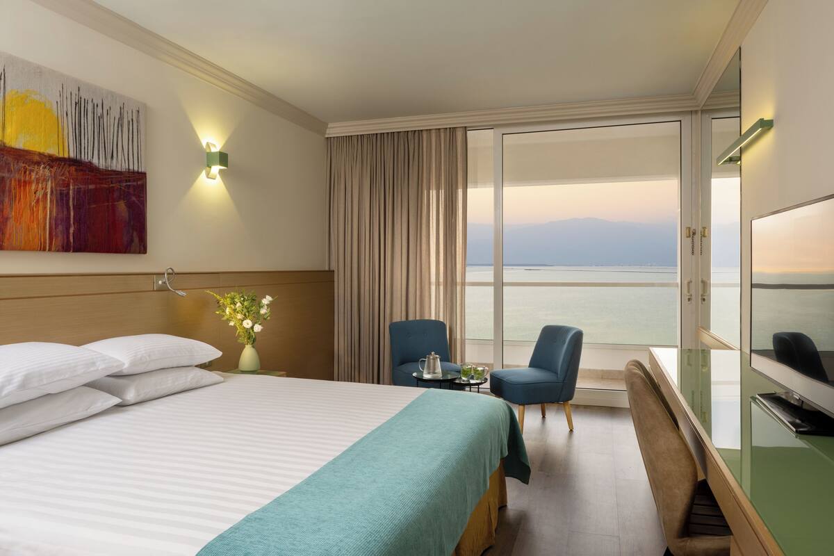 A hotel room with a bed, a desk, two chairs, a TV, and a large window with a view of the sea.