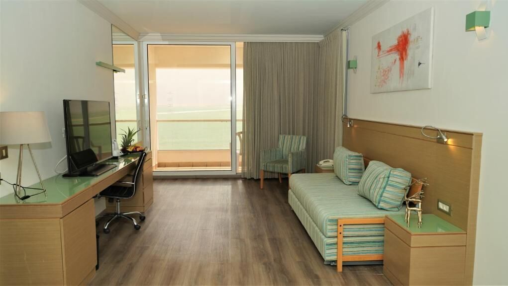 Habitación de hotel con cama, escritorio con silla, televisor, lámpara y vista al balcón.