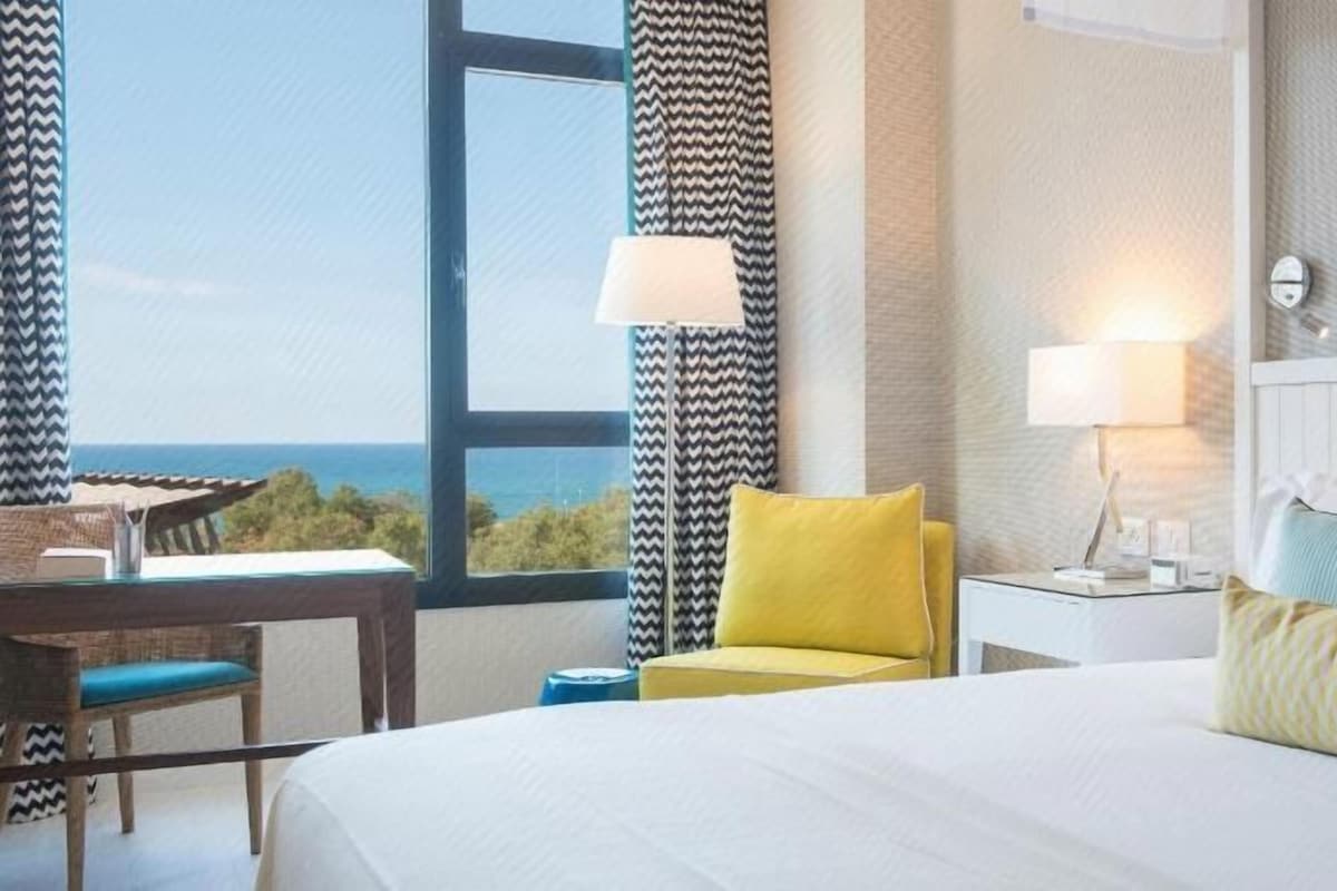 A hotel room with a bed, a desk, a chair, and a view of the sea.