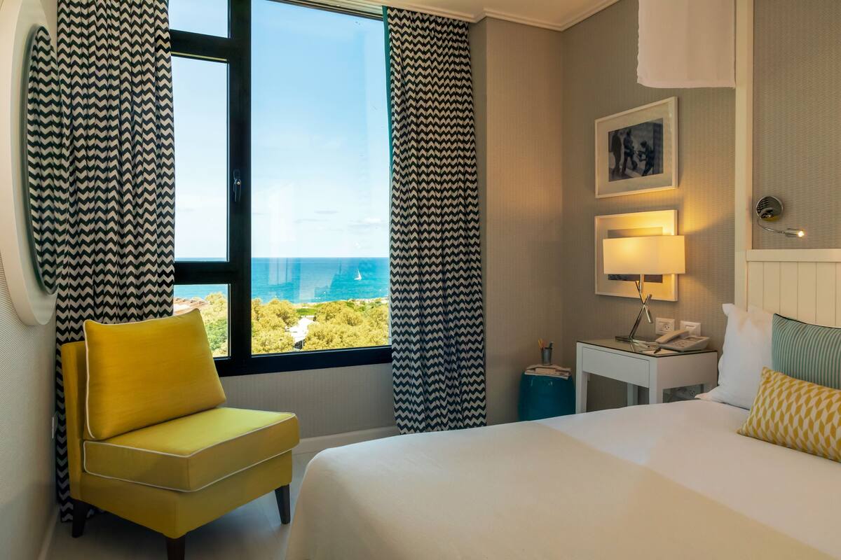 Une chambre d’hôtel avec un lit, une chaise, un bureau avec une lampe et une vue sur l’océan.