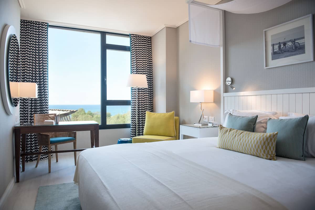 Une chambre d’hôtel avec un grand lit, un bureau avec une chaise, une petite table et une fenêtre avec des rideaux.