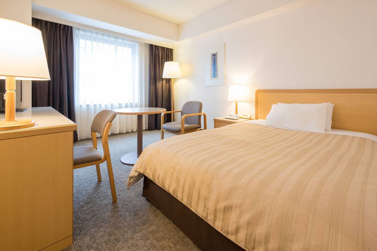 Habitación de hotel con cama, dos sillas, escritorio, lámpara, mesita de noche y ventana con cortinas.