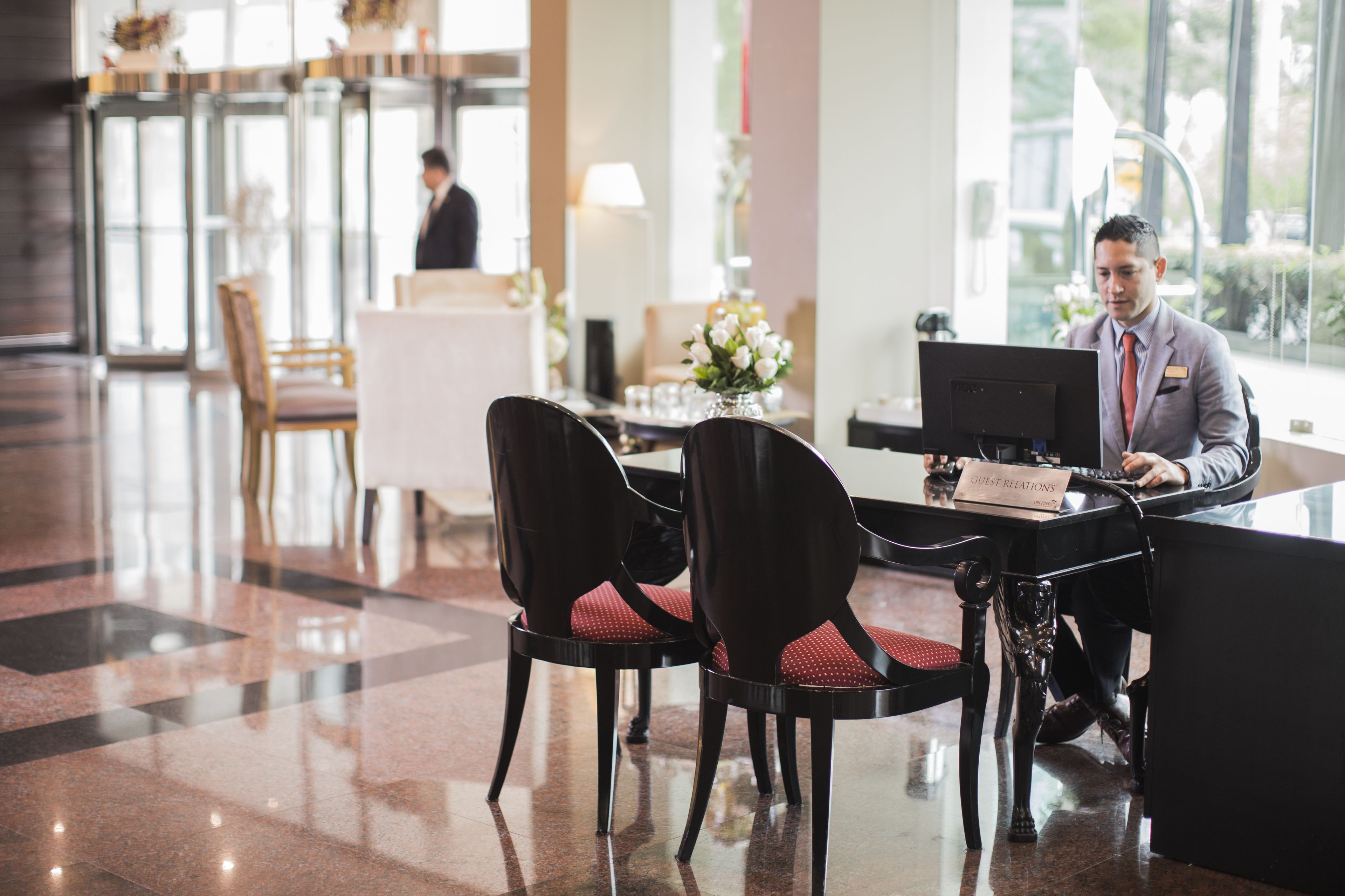 Zona de recepción de hotel con una recepcionista en el mostrador, dos sillas negras con cojines rojos y una placa que dice “Guest Relations”.