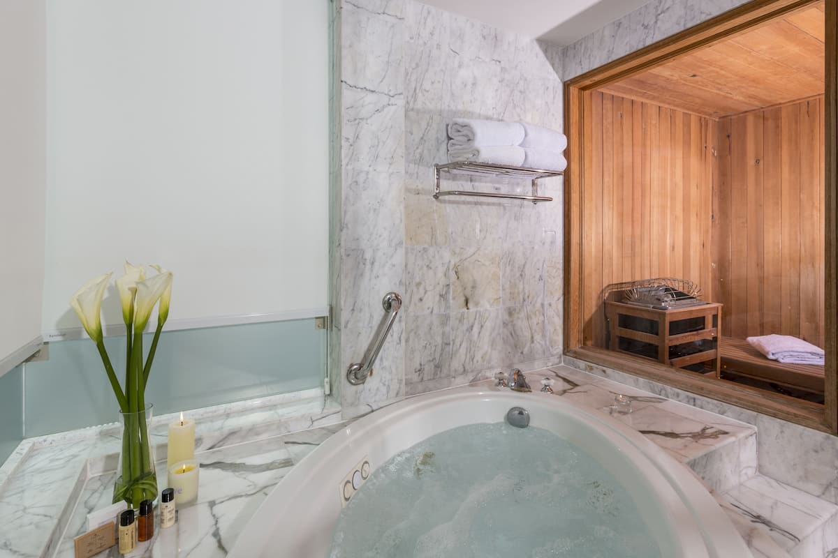 Um banheiro com uma banheira de mármore, uma sauna de madeira, um vaso de vidro com flores e um porta-toalhas.