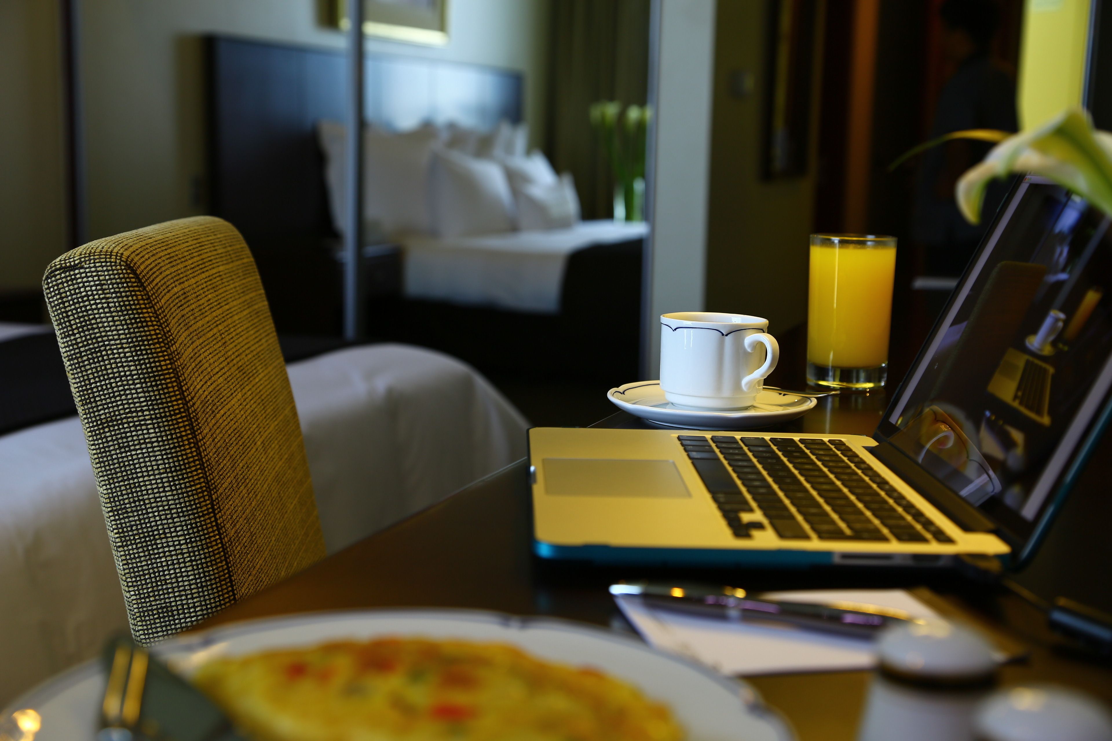 Una habitación de hotel con una cama, un ordenador portátil sobre una mesa, una taza de café, un vaso de zumo de naranja y un plato de comida.
