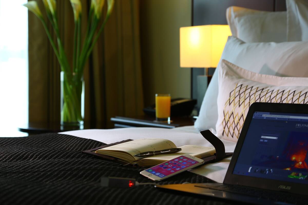 Quarto de hotel com uma cama, um laptop, um smartphone, um caderno e uma caneta sobre a mesa de cabeceira.