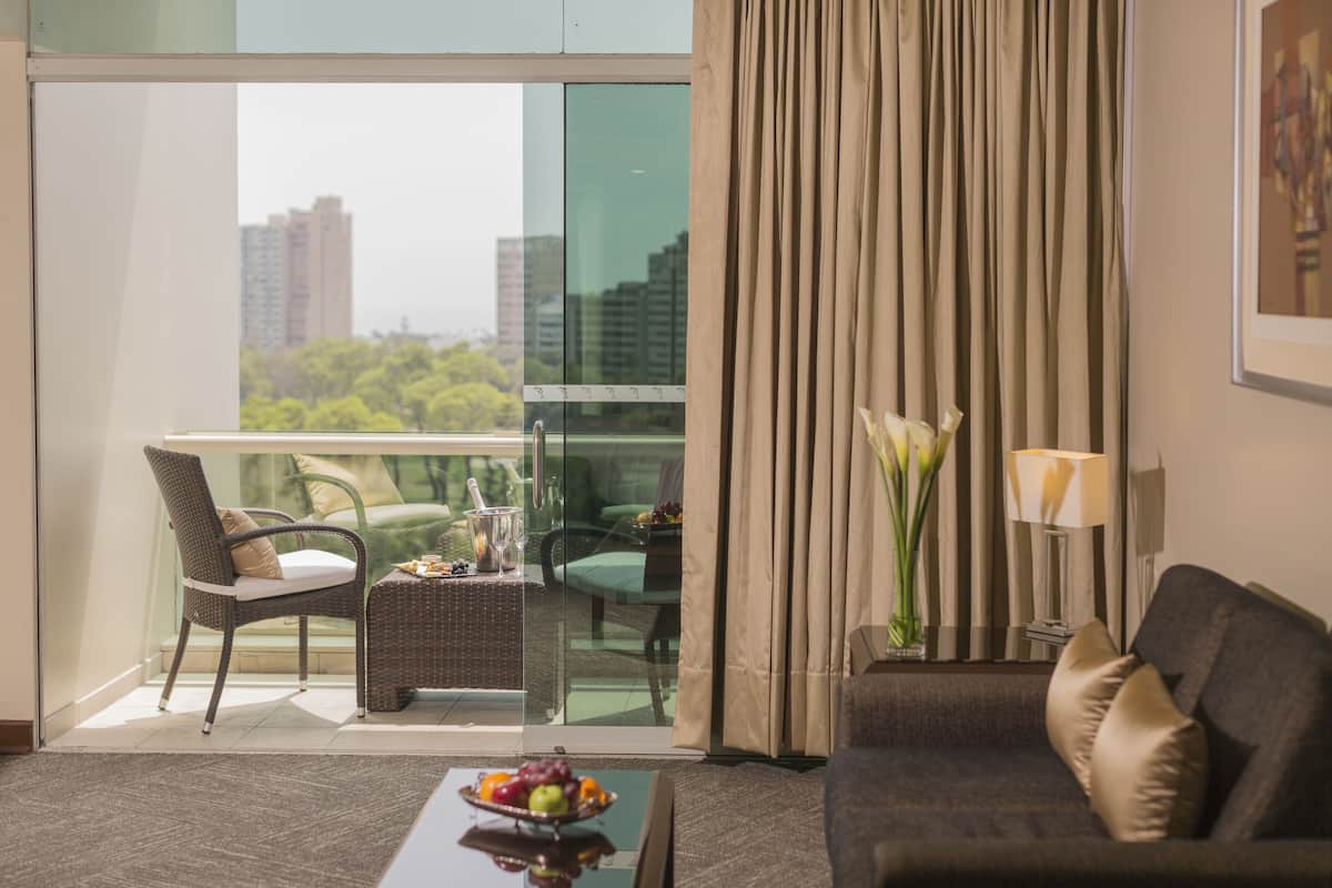 Quarto de hotel com varanda, área de refeições com mesa e cadeiras, sofá e um vaso com flores.
