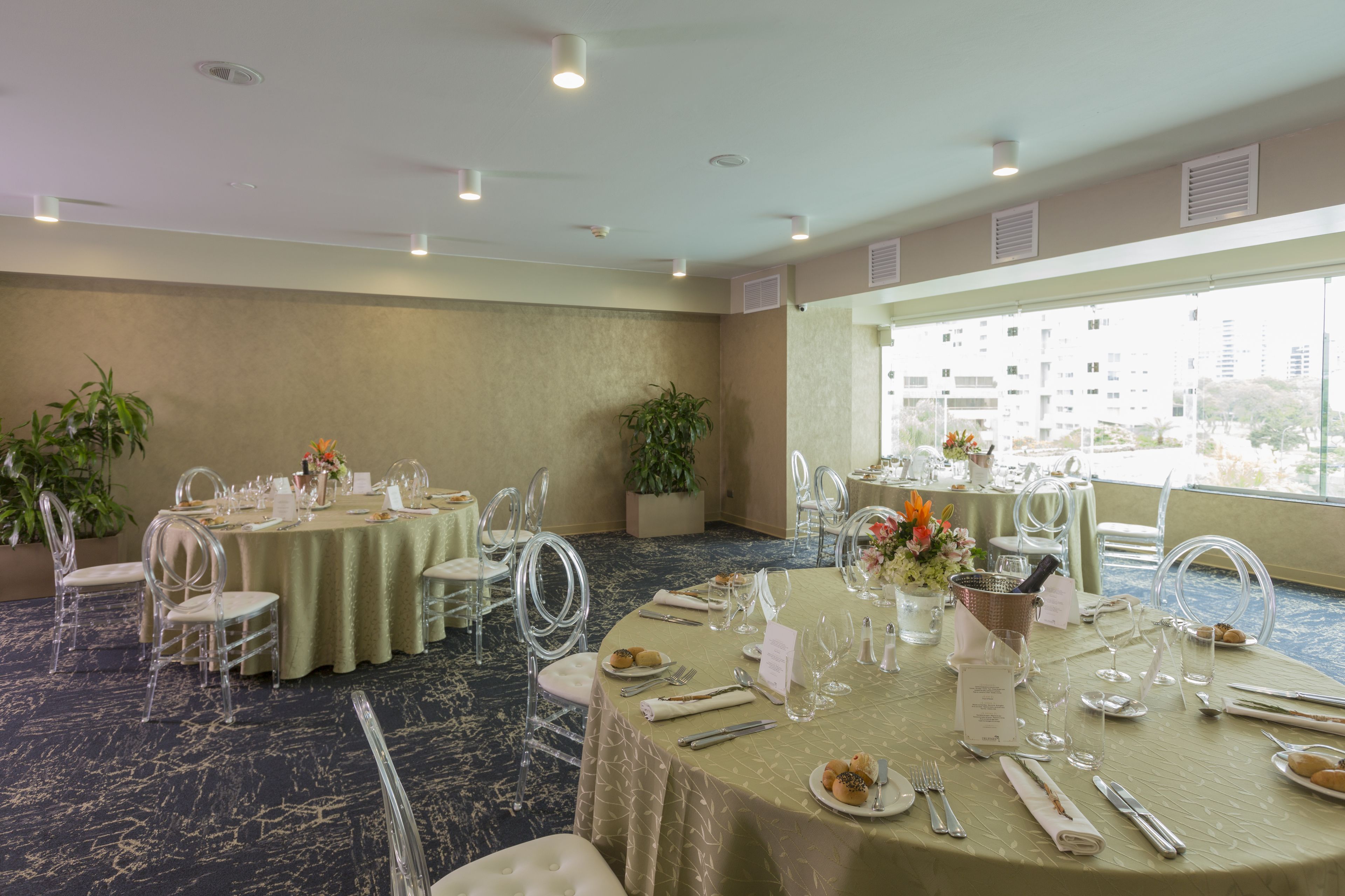A banquet hall with round tables set for dining, clear chairs, and potted plants.