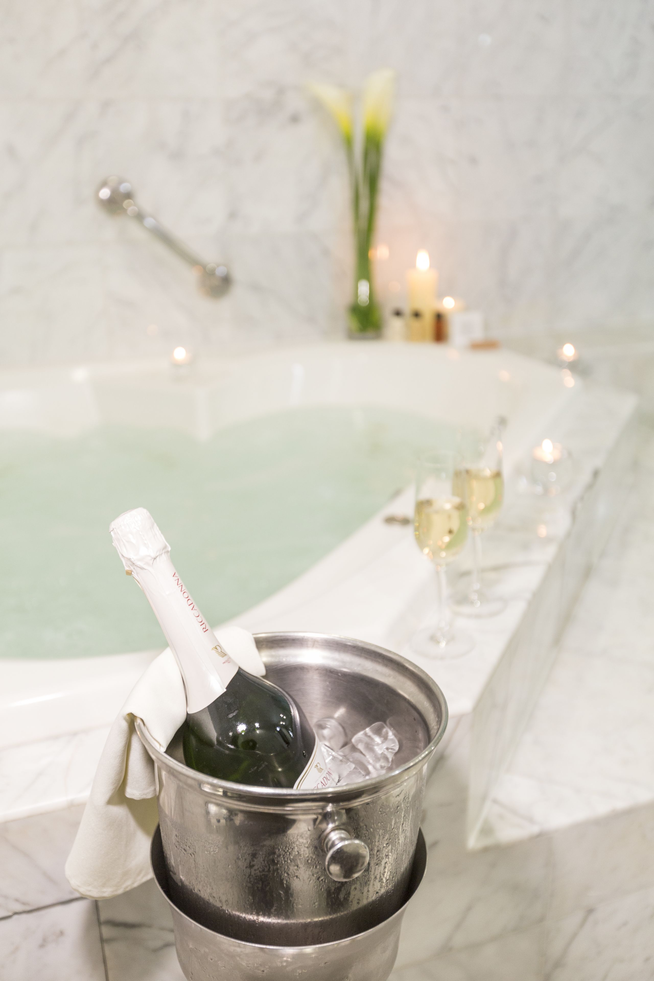Una bañera con champán, copas, una toalla y velas en un baño con pared de mármol.