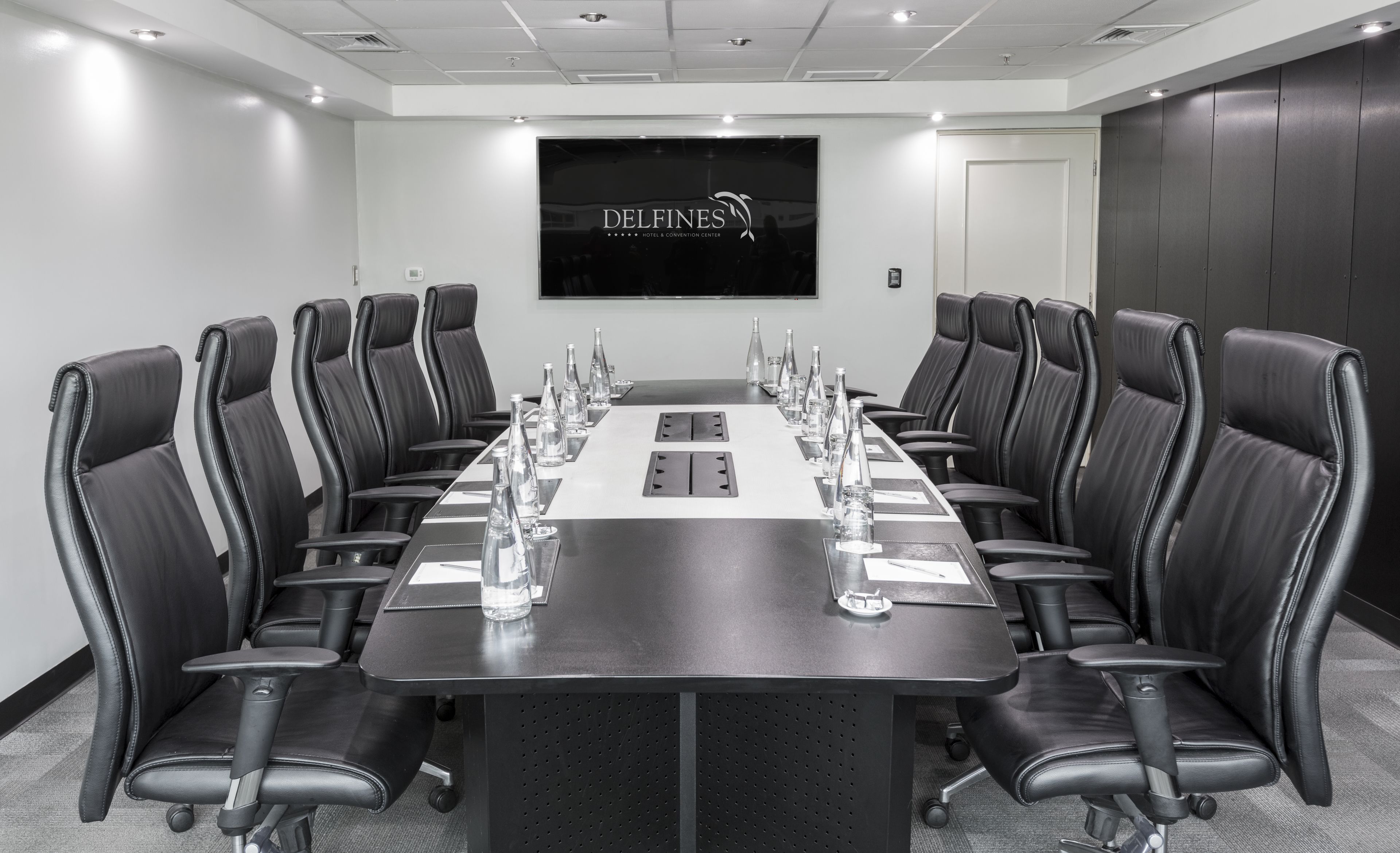 Un salón de conferencias con una larga mesa, sillas de cuero negro y una pantalla grande que muestra 'DELFINES HOTEL & CONVENTION CENTER'.