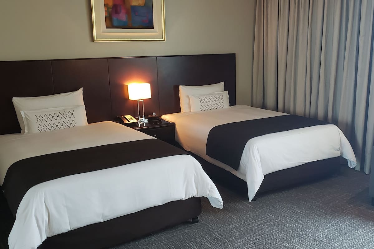 Quarto de hotel com duas camas, uma mesa de cabeceira com abajur, uma cadeira e um quadro na parede.