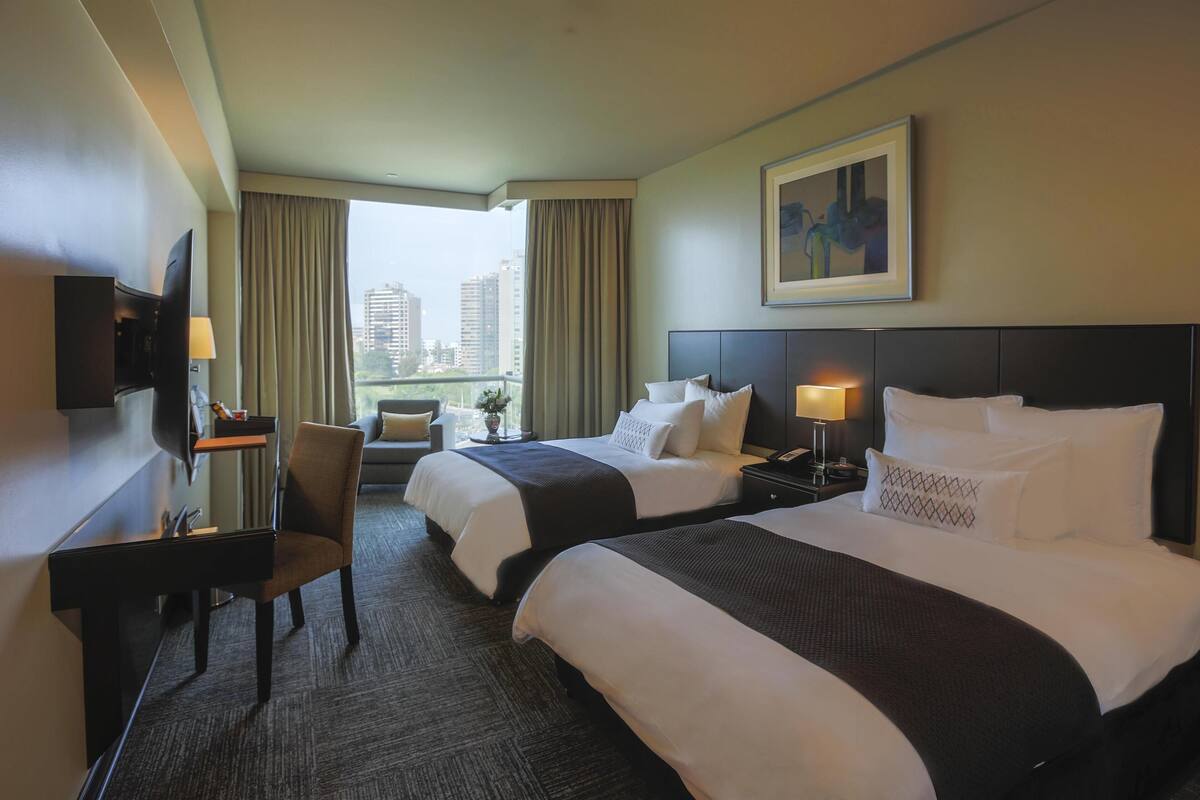 Quarto de hotel com duas camas, uma escrivaninha, uma cadeira, televisão e vista para a cidade.