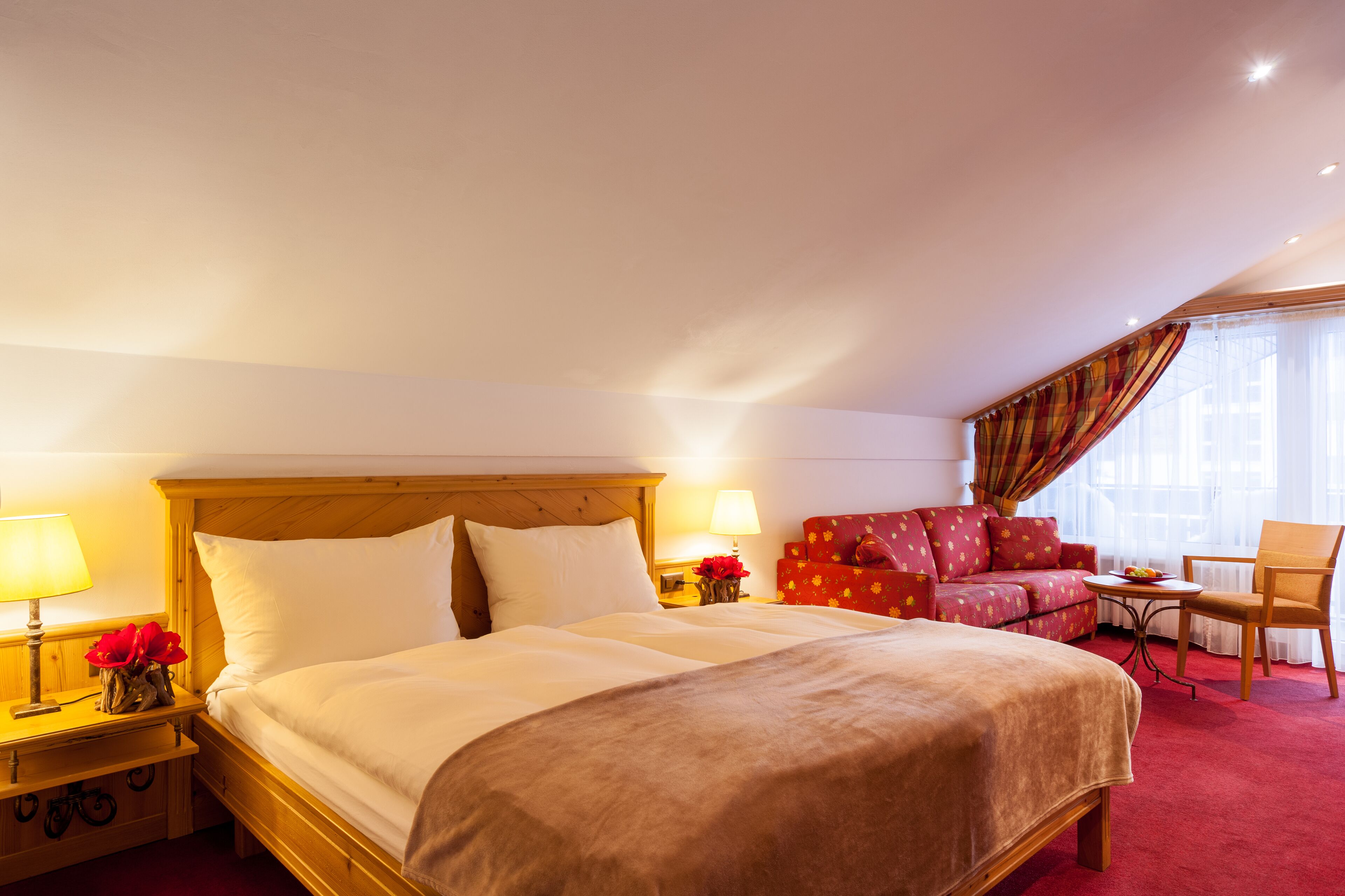 A hotel room with a large bed, a red sofa, a small table, and a chair.