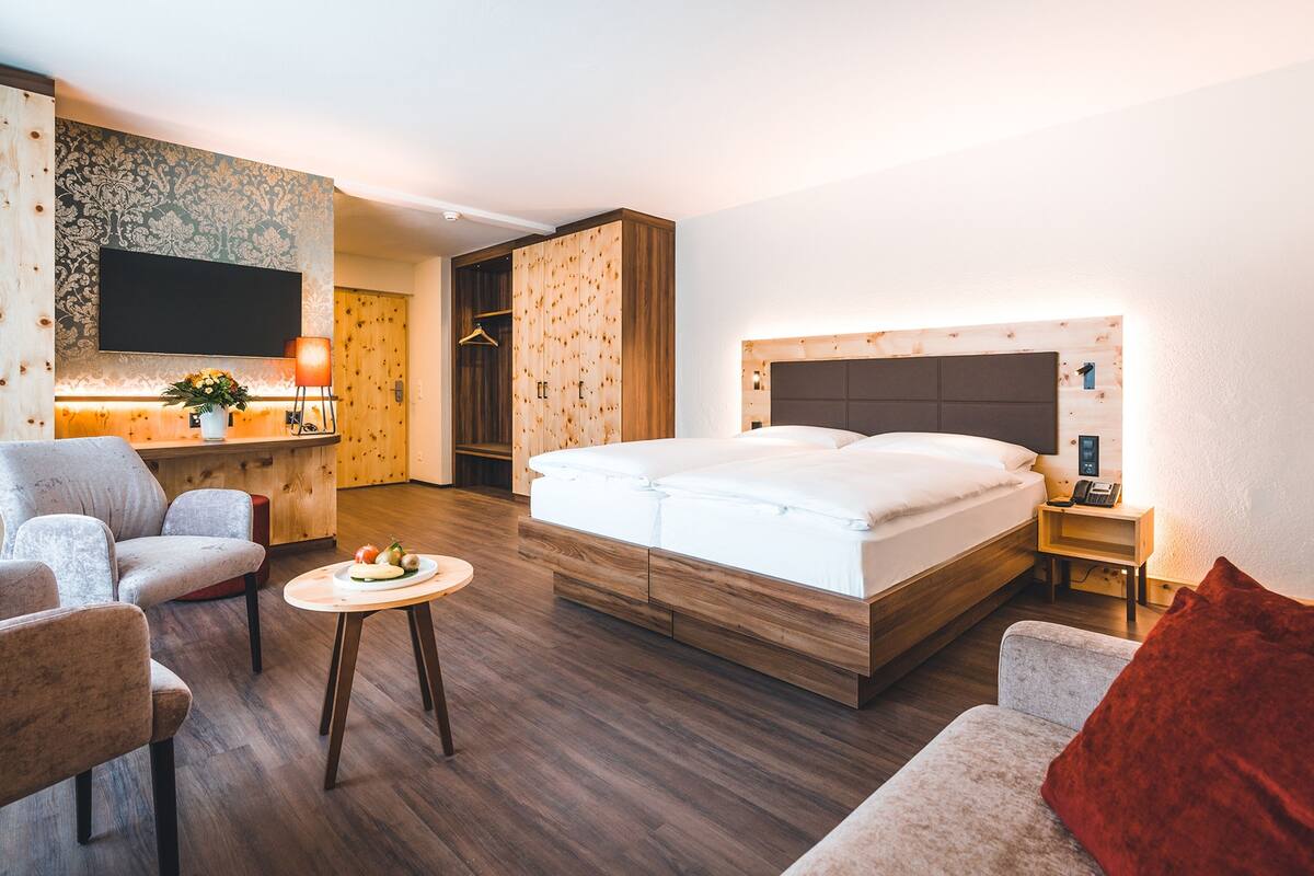 Une chambre d’hôtel moderne avec un grand lit, un coin salon comprenant un canapé et un fauteuil, une petite table ornée d’un vase de fleurs et une armoire en bois.