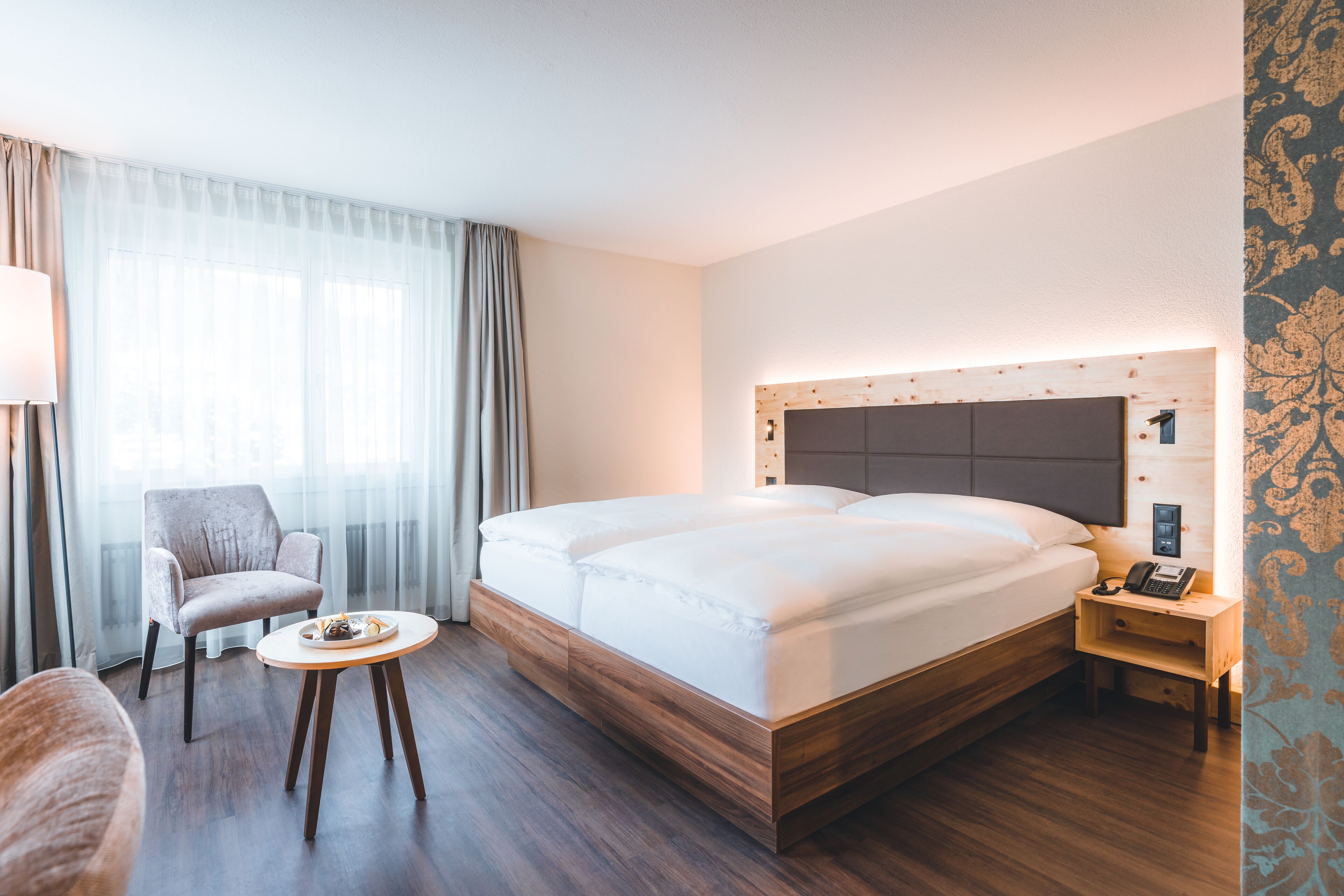 A modern hotel room with a large bed, a chair, a small table, and a nightstand with a phone.