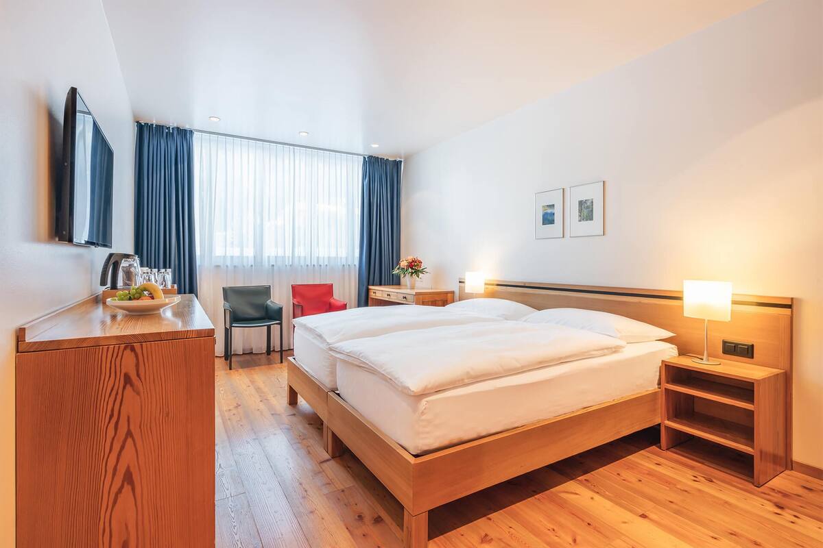 A hotel room with a wooden bed, a desk with a bowl of fruit, a chair, a TV, and a window with curtains.