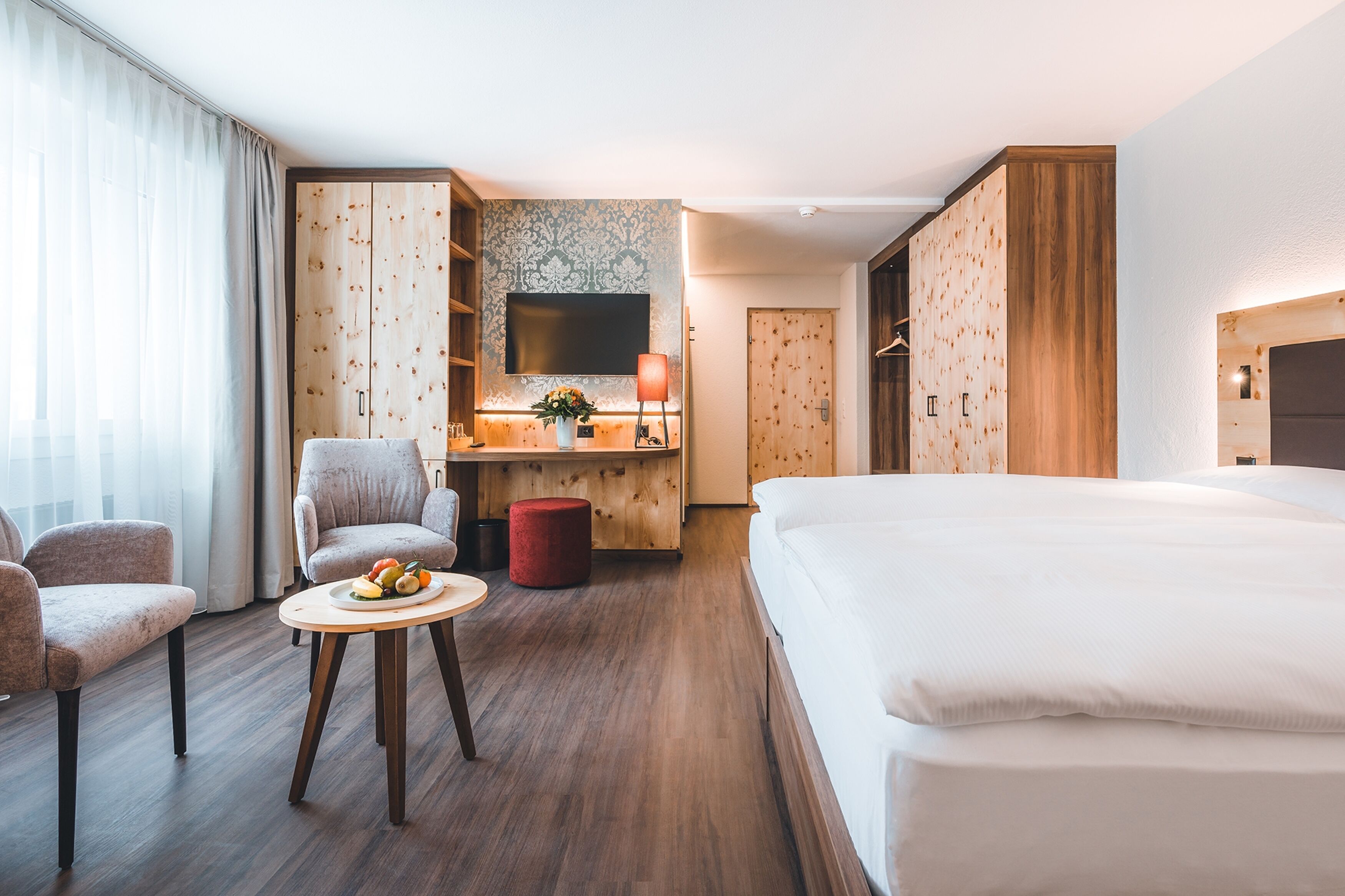 Een hotelkamer met een groot bed, een bureau met een televisie, een stoel, een klein tafeltje met fruit, en een rode poef.