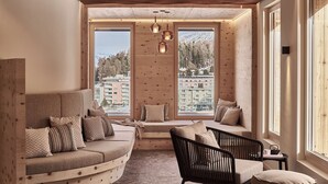 Una habitación acogedora con un sofá curvo, una silla negra y una pequeña mesa. Amplias ventanas ofrecen vistas a edificios y árboles.