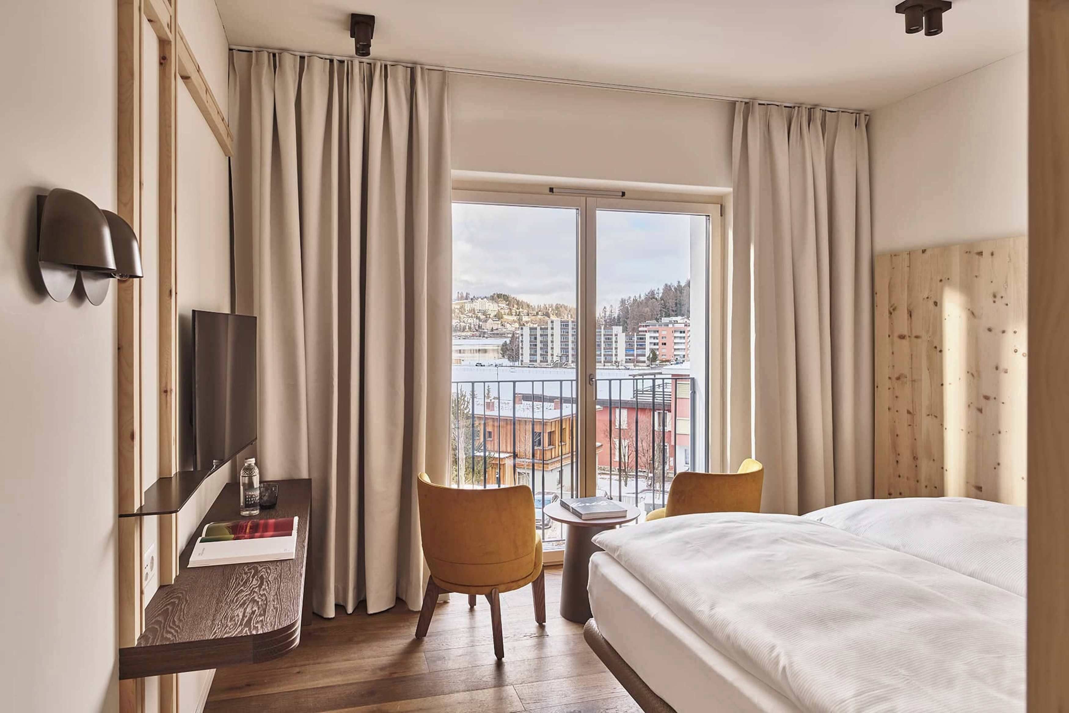 A hotel room with a bed, a desk with a chair, a TV, and a view of the city through large windows.