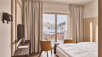 A hotel room with a bed, a desk with a chair, a TV, and a view of the city through large windows.