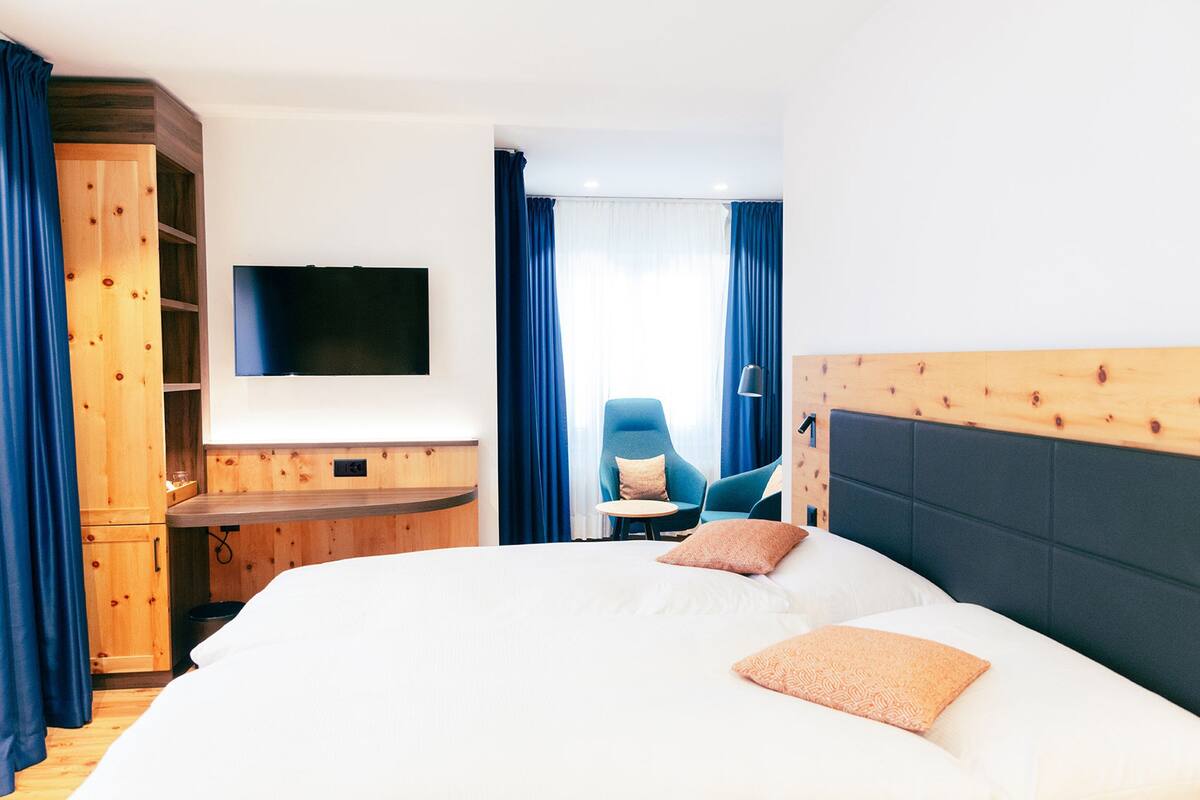 A hotel room with a large bed, a TV mounted on the wall, a wooden cabinet, and a seating area with a chair and table.