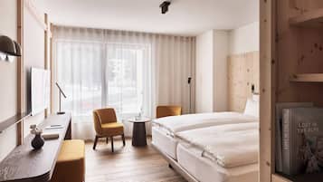 Een hotelkamer met een bed, een bureau met stoel, een kleine tafel met stoel, een boekenplank en een groot raam met gordijnen.