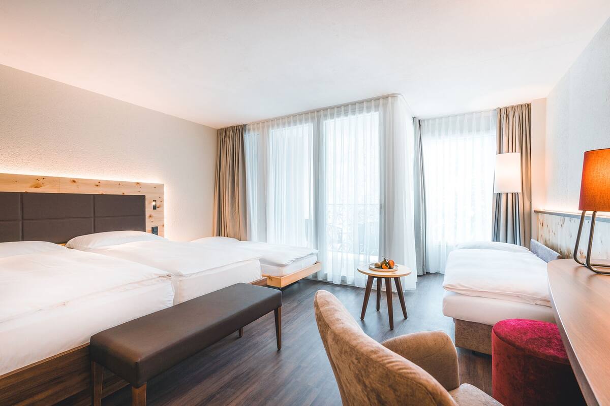 Une chambre d’hôtel avec deux lits, un banc, une petite table avec des fruits et une lampe.