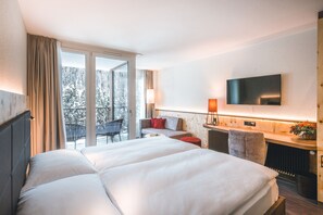 A hotel room with a large bed, a desk with a chair, a TV mounted on the wall, and a balcony with a view of trees. - Hotel Laudinella (St. Moritz)