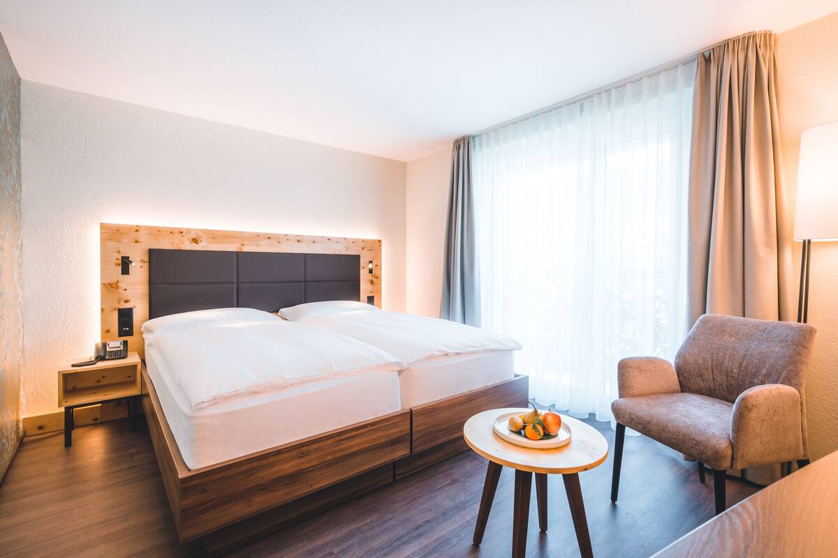 Une chambre d’hôtel avec un lit en bois, une table de chevet avec une lampe, une chaise et une petite table sur laquelle se trouvent des fruits.