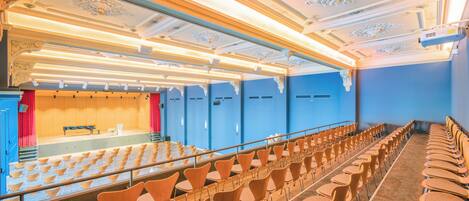 Un auditorio con hileras de sillas, un escenario y detalles ornamentales en el techo.