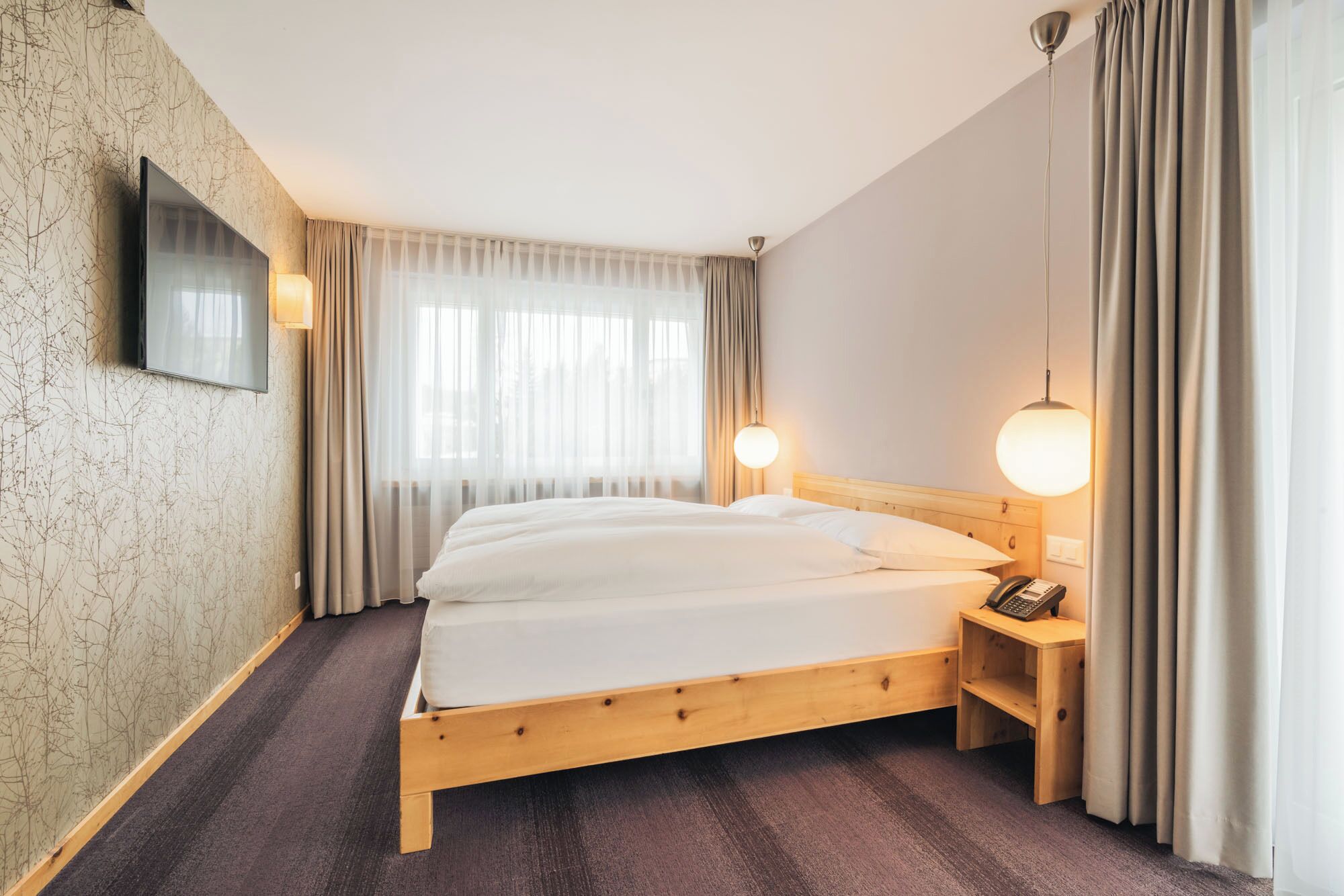 Une chambre d’hôtel avec un lit en bois, un téléviseur à écran plat fixé au mur, une table de chevet avec une lampe et une fenêtre ornée de rideaux légers.