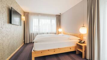 Une chambre d’hôtel avec un lit en bois, un téléviseur à écran plat fixé au mur, une table de chevet avec une lampe et une fenêtre ornée de rideaux légers.