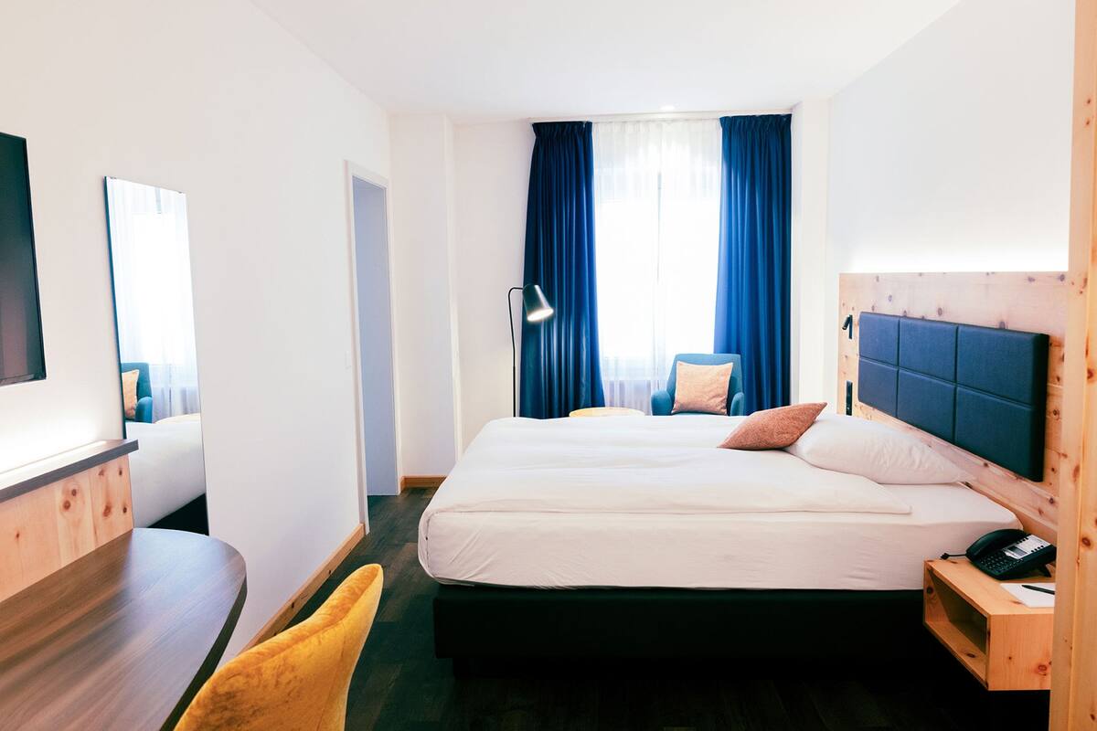 A hotel room with a large bed, a desk, a chair, and a bedside table with a lamp.