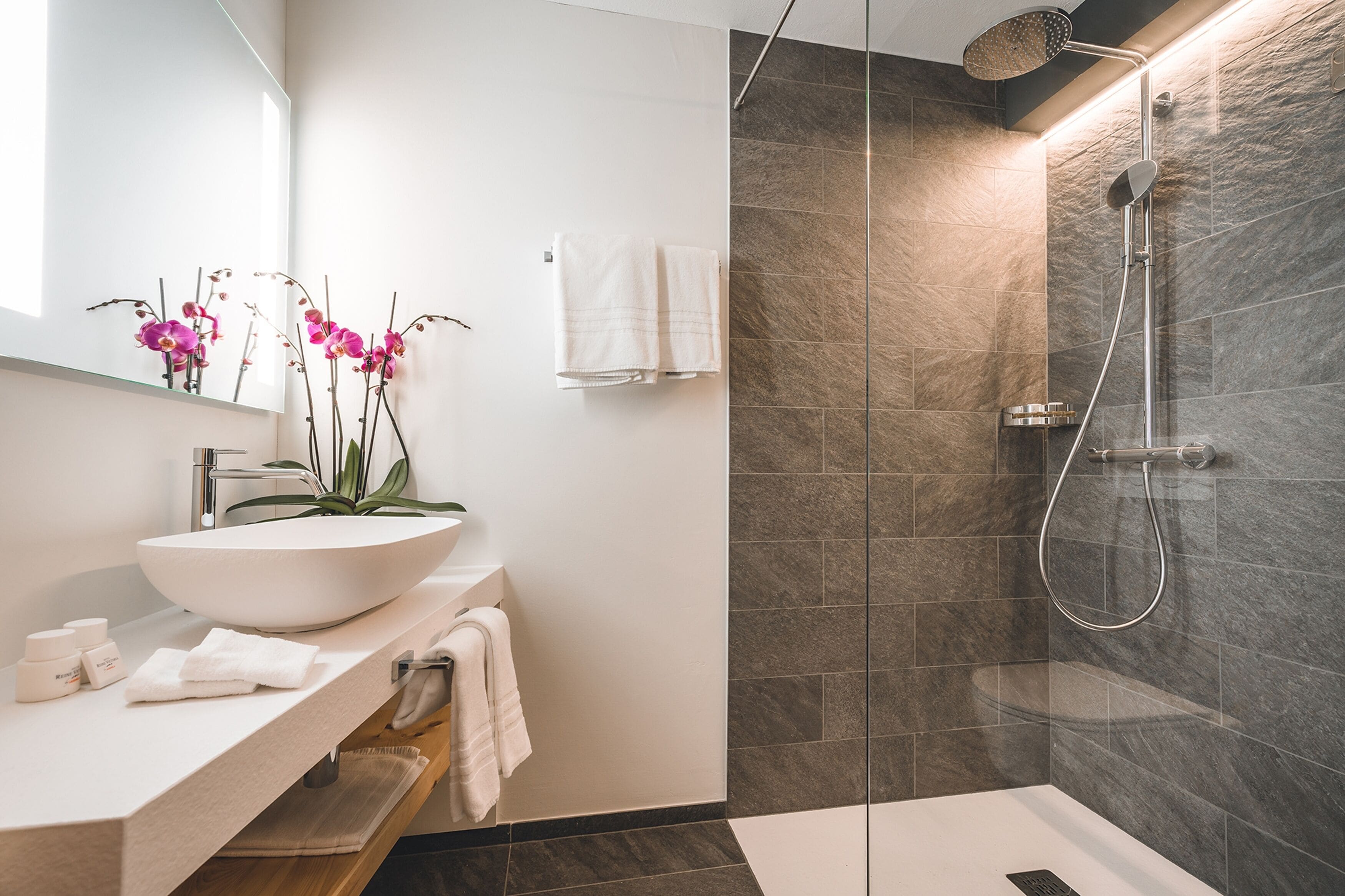 A modern bathroom with a large white sink, a mirror, a shower with a glass door, and a vase with pink orchids.