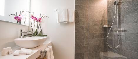 A modern bathroom with a large white sink, a mirror, a shower with a glass door, and a vase with pink orchids.