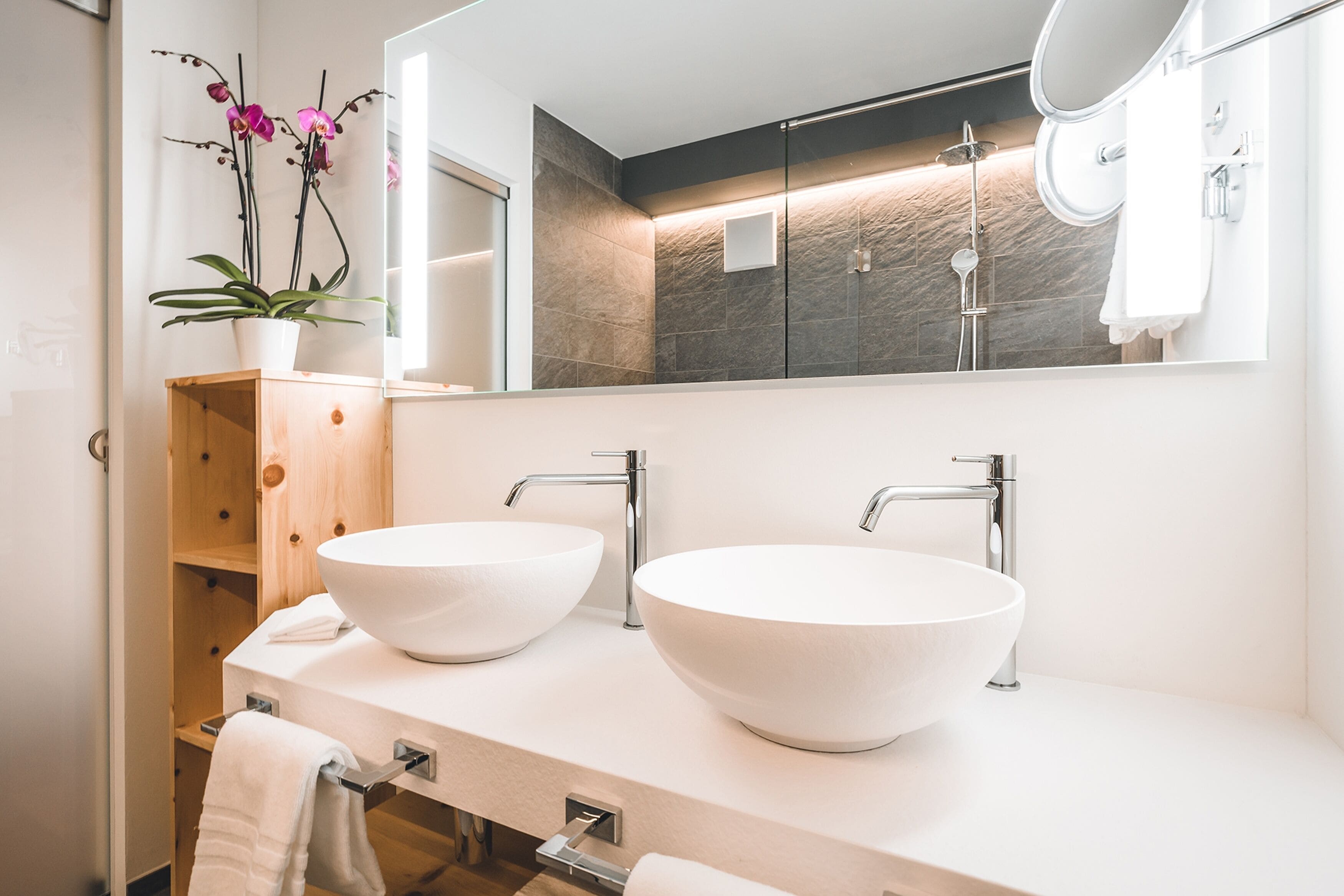 A modern bathroom with two large white sinks, a wooden cabinet, a potted orchid, and a large mirror.