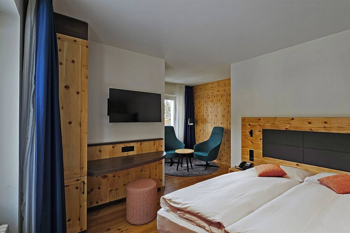 A hotel room with a bed, a TV mounted on the wall, a wooden headboard, a desk with a chair, and a window with curtains.