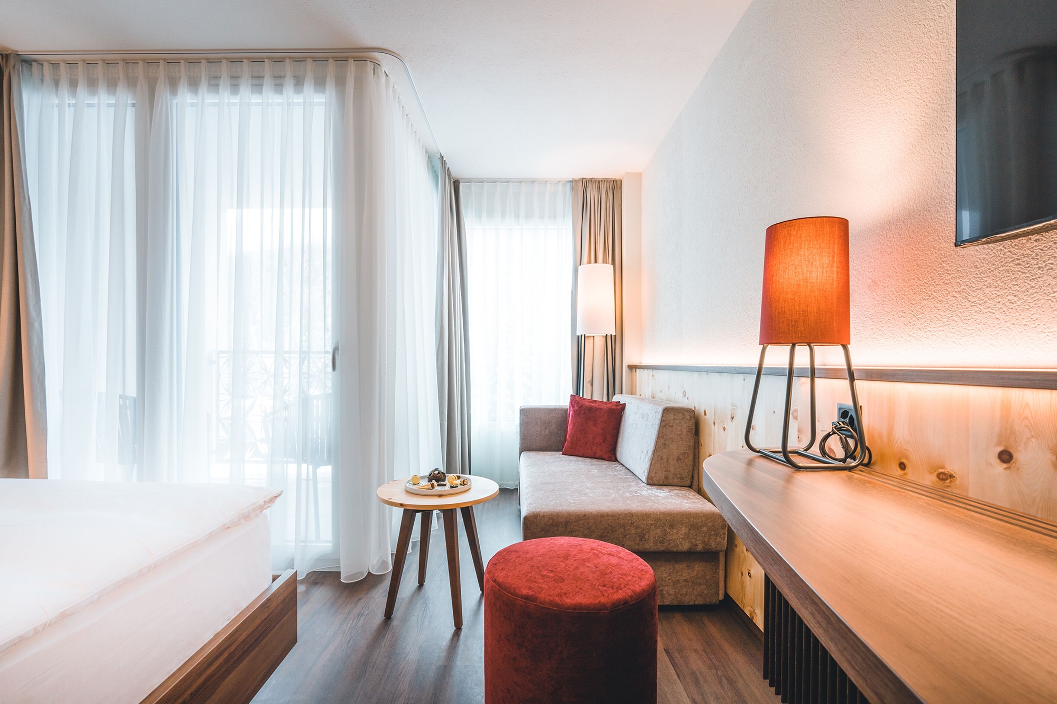 Een hotelkamer met een bed, een sofa, een rode poef, een klein tafeltje met een schaal lekkernijen, een lamp en een aan de muur bevestigde televisie.