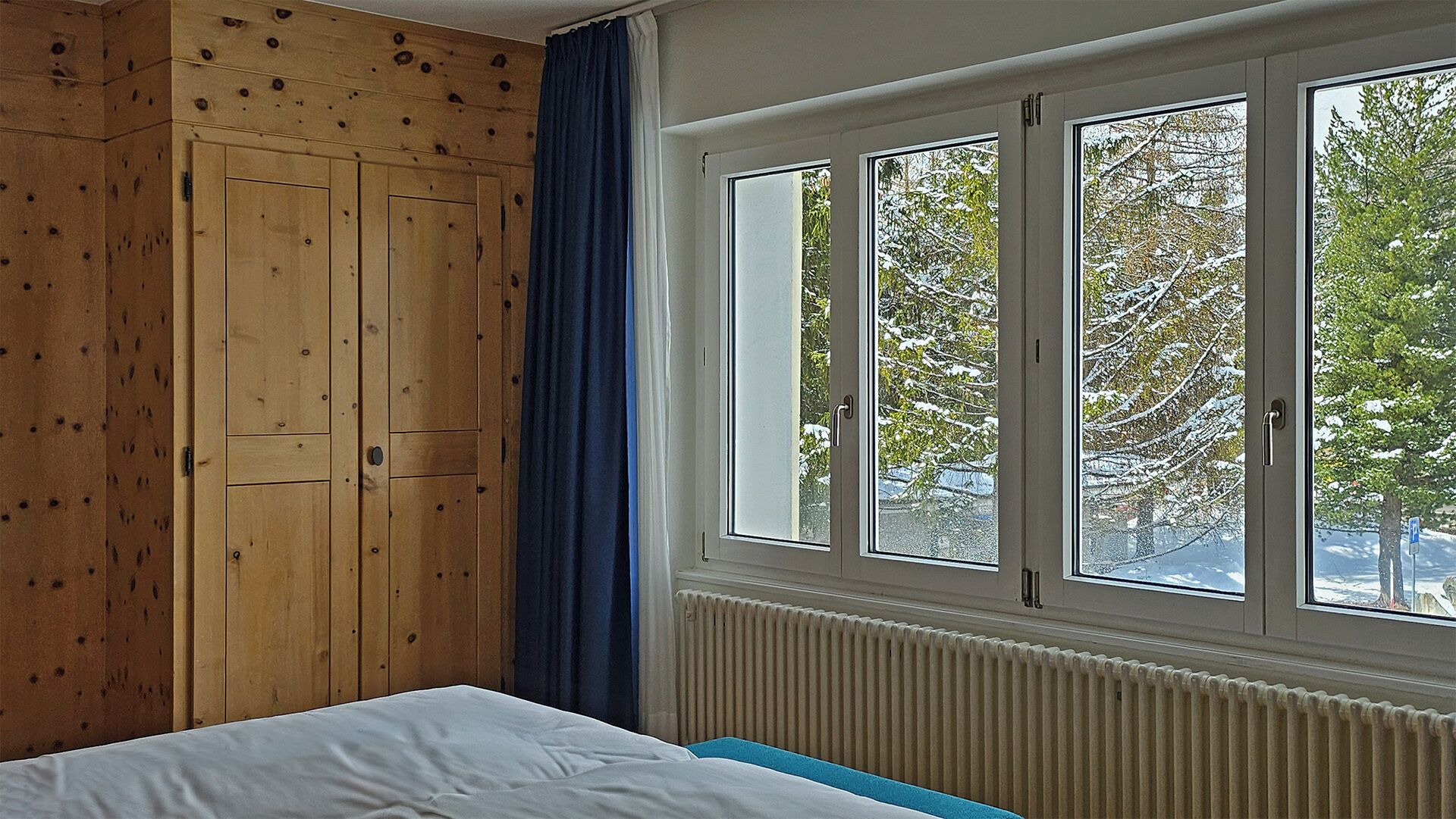 Un dormitorio con un armario de madera, una cama, un radiador y una ventana con vistas a árboles.