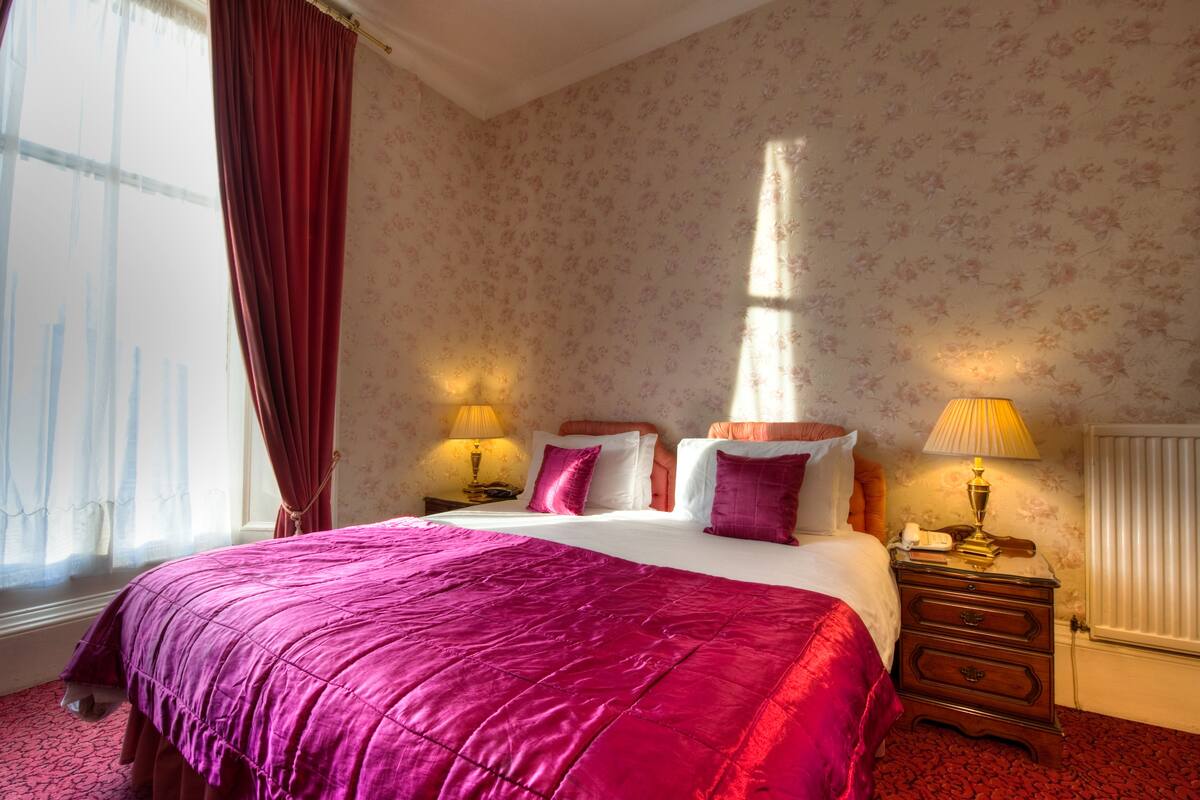A hotel room with a large bed, floral wallpaper, and two bedside lamps.