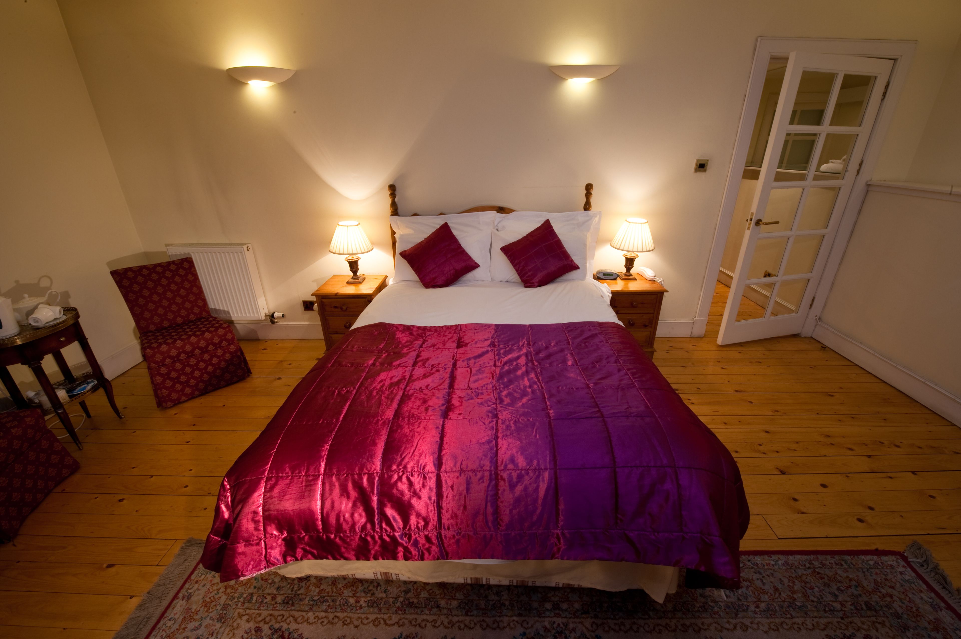 Ein Schlafzimmer mit einem Bett, zwei Nachttischlampen, einem roten Sessel, einem Heizkörper und einer Tür zu einem anderen Raum.